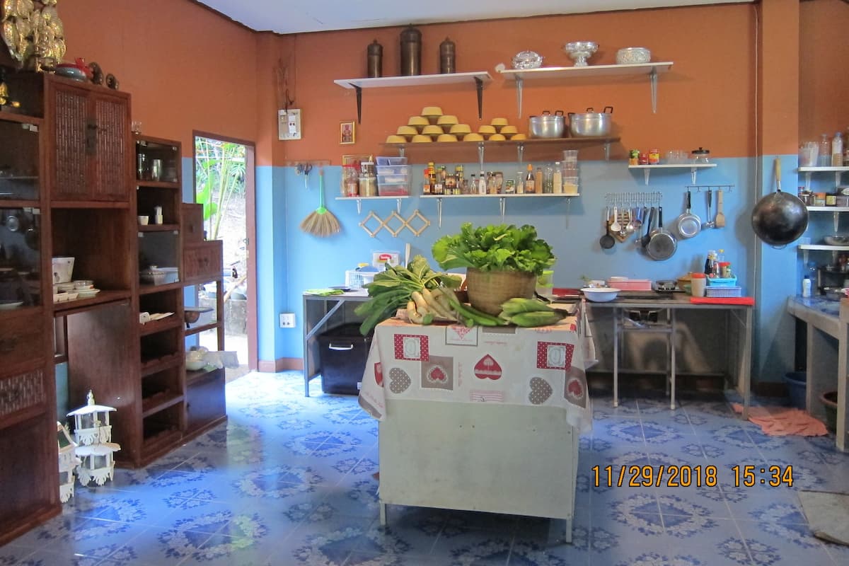 Une cuisine avec une table centrale recouverte d’une nappe, entourée d’étagères remplies d’ustensiles de cuisine et un balai accroché au mur.