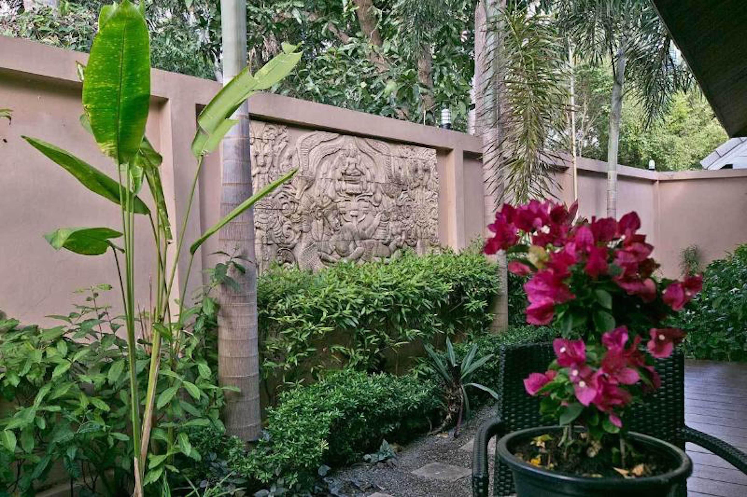 A garden with a stone wall featuring carvings, lush green plants, and a potted plant with pink flowers.