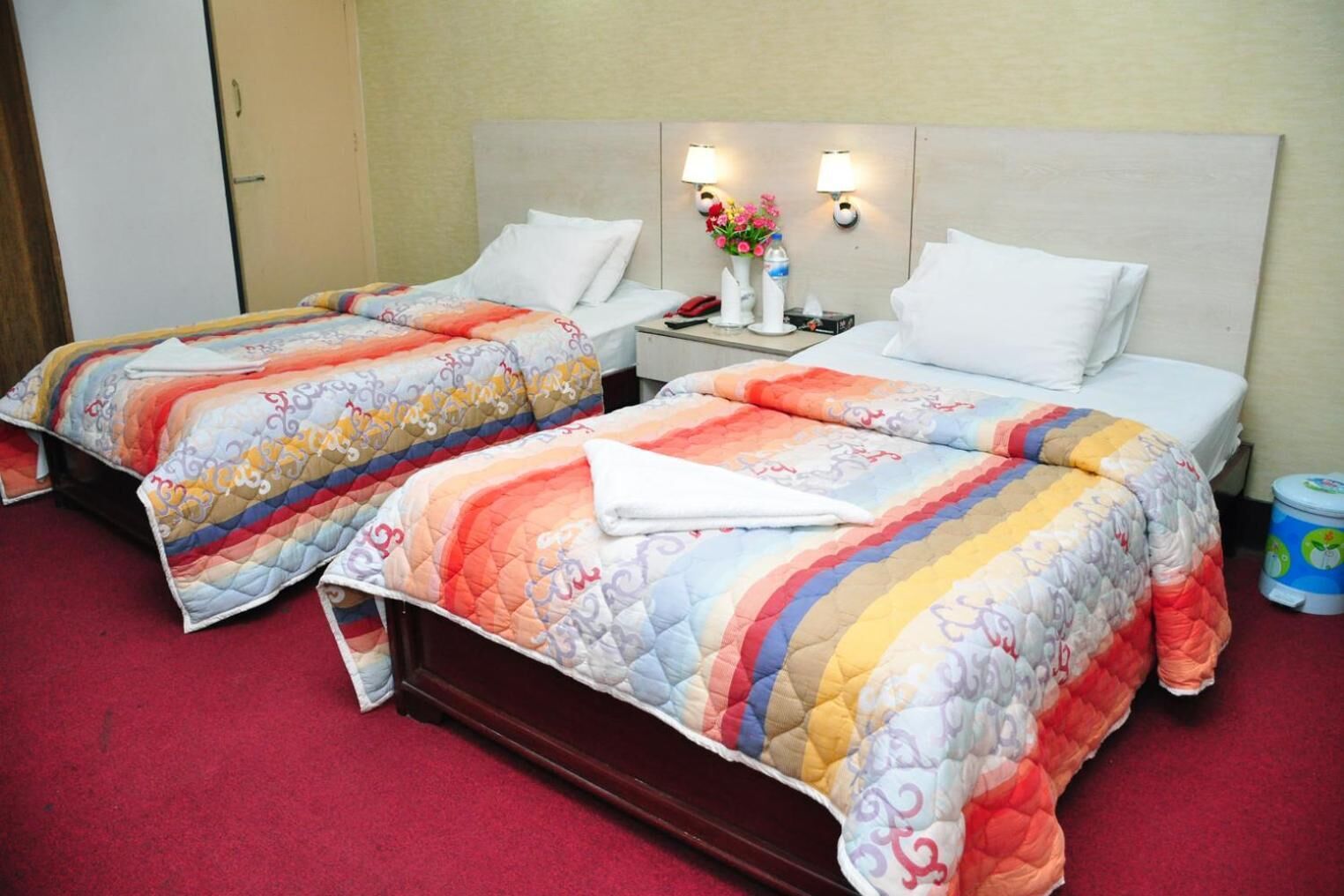 A hotel room with two beds, each with a colorful quilt, a nightstand with a lamp, and a vase with flowers.