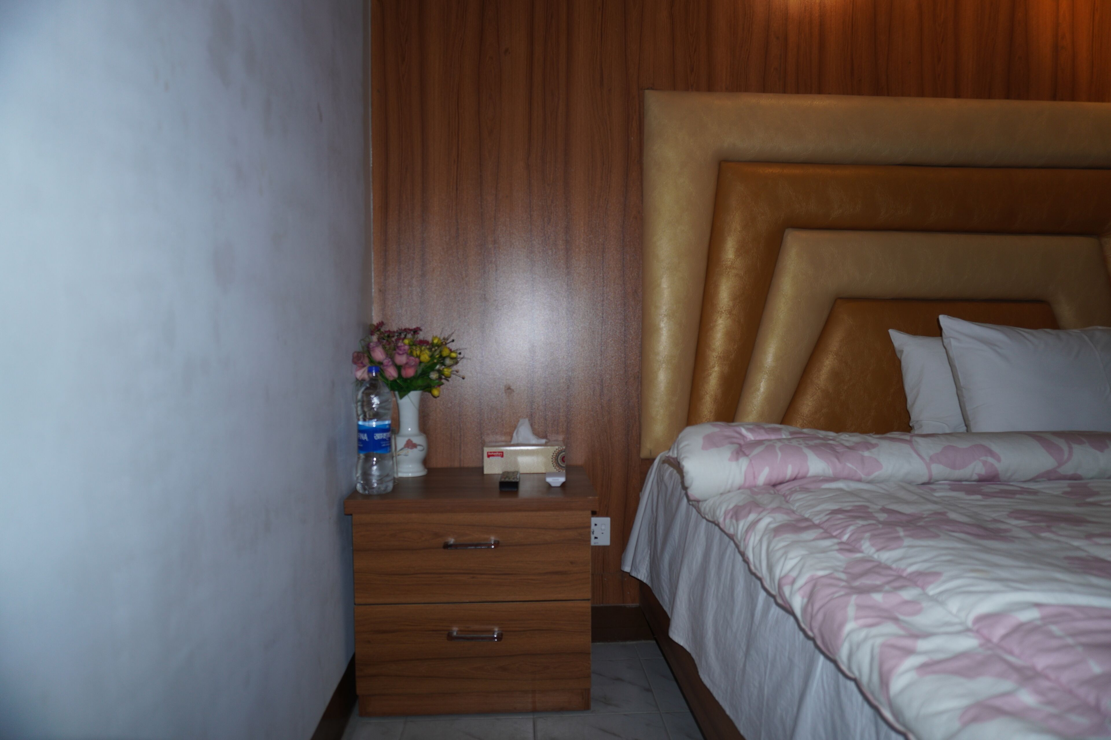 Une chambre d’hôtel avec une tête de lit en bois, une table de chevet ornée d’un vase de fleurs et un lit recouvert d’une couvre-lit à motifs.