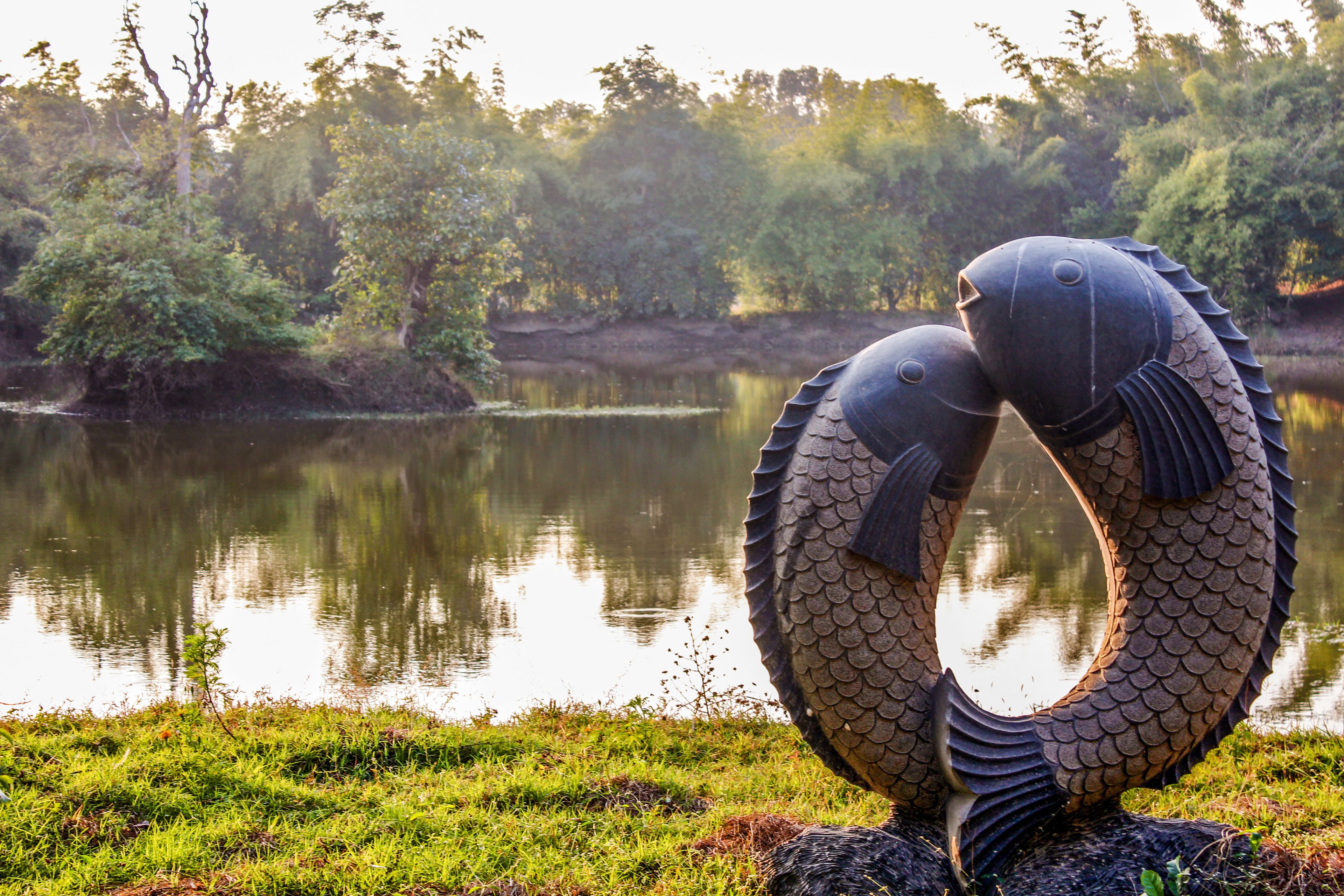 A sculpture of two fish intertwined in a circular formation, set against a calm lake with trees in the background.