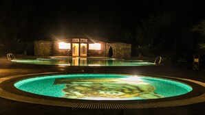 A nighttime swimming pool with a circular design, a stone building in the background, and a well-lit entrance. - Syna Tiger Resort - Bandhavgarh (Manpur)