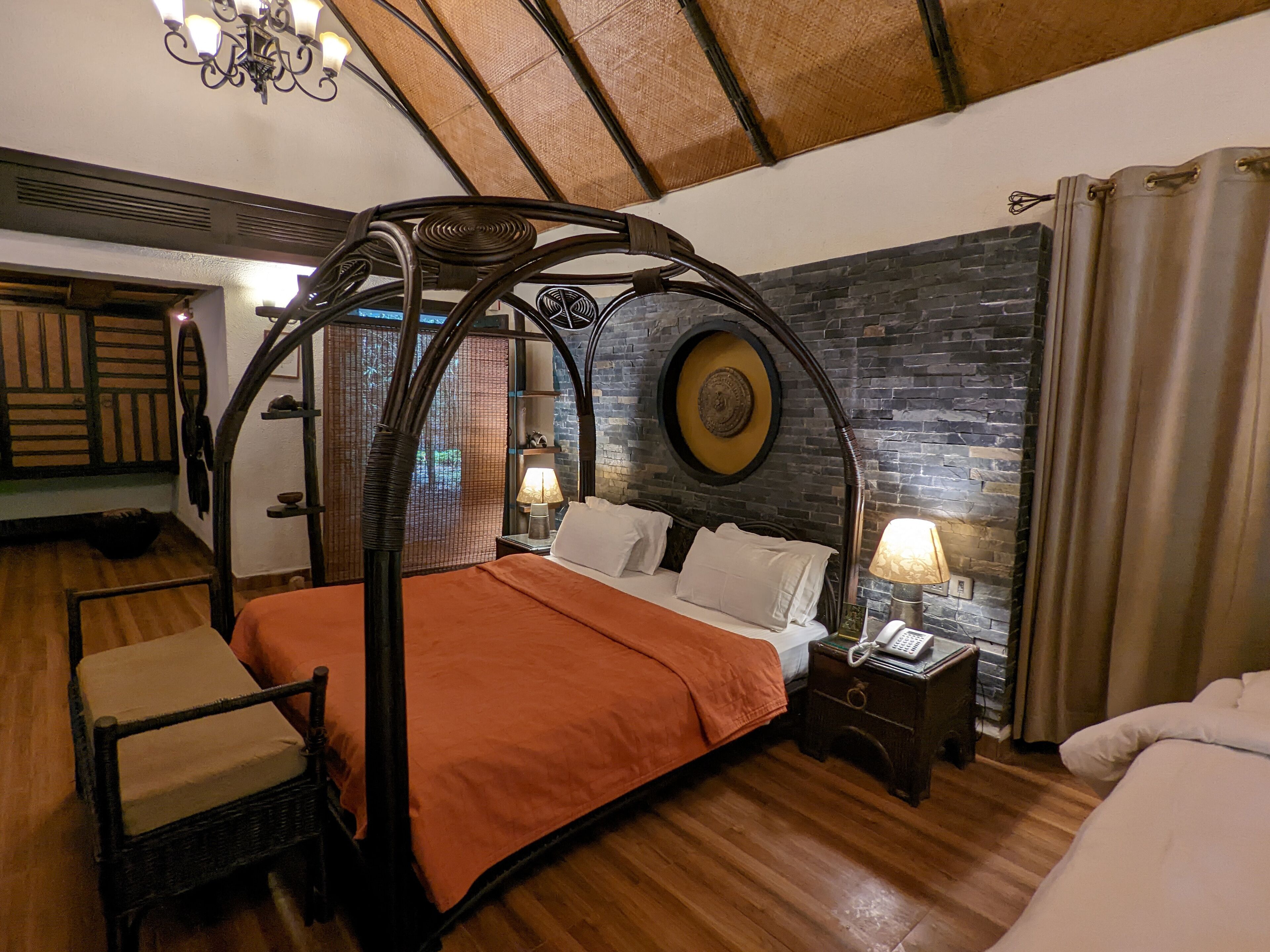 A four-poster bed with an orange bedspread, a nightstand with a lamp, and a stone wall with a circular design.
