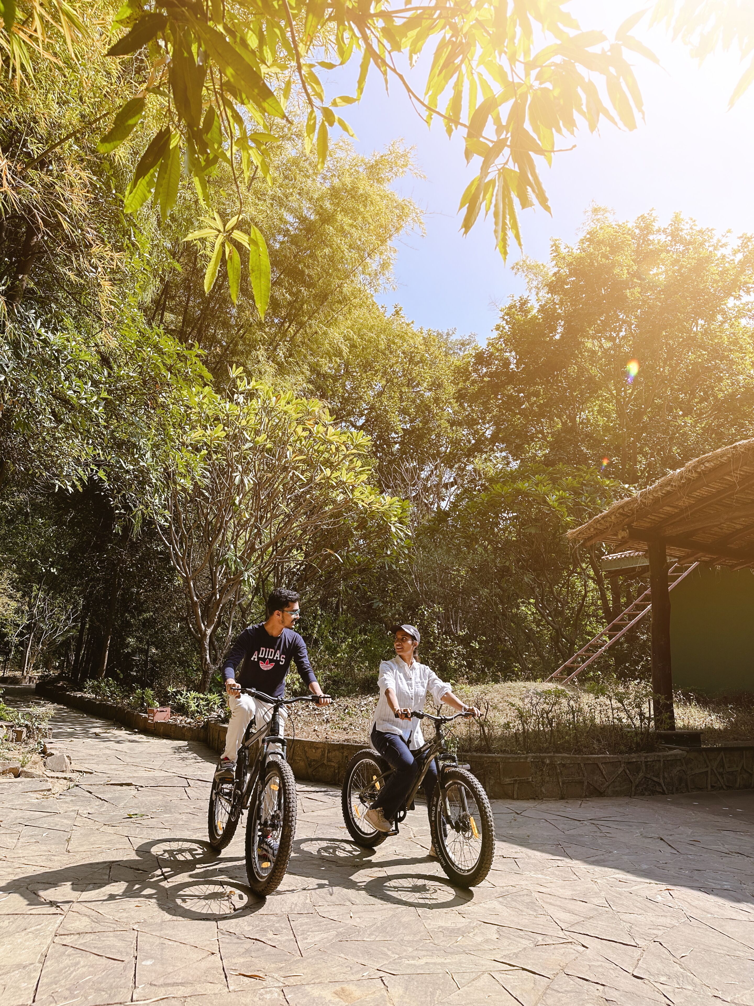 Two people on bicycles in a paved outdoor area with trees and a structure in the background.