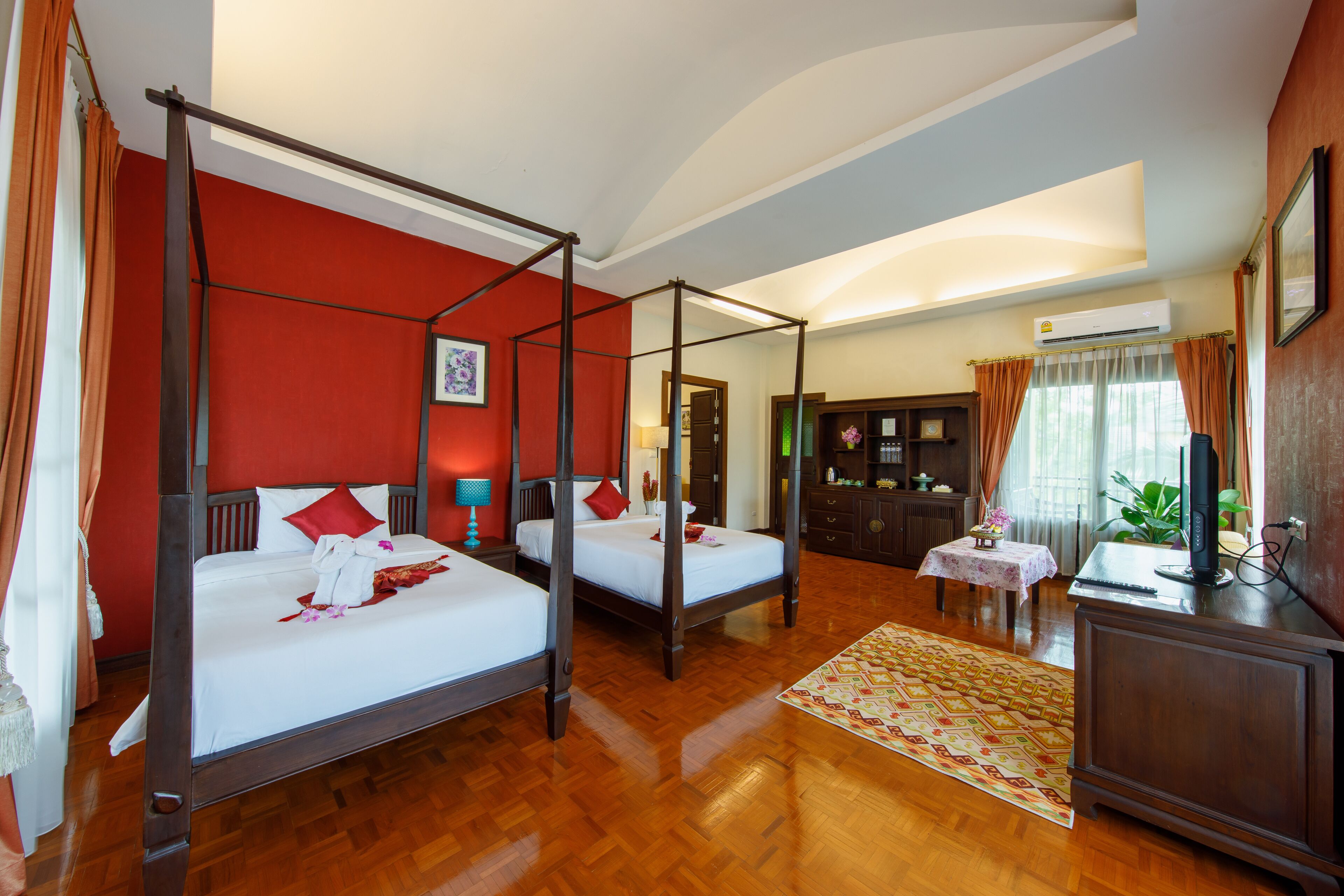 A four-poster bed with white linens and red pillows, a wooden floor, a small table with a floral tablecloth, and a television on a wooden cabinet.