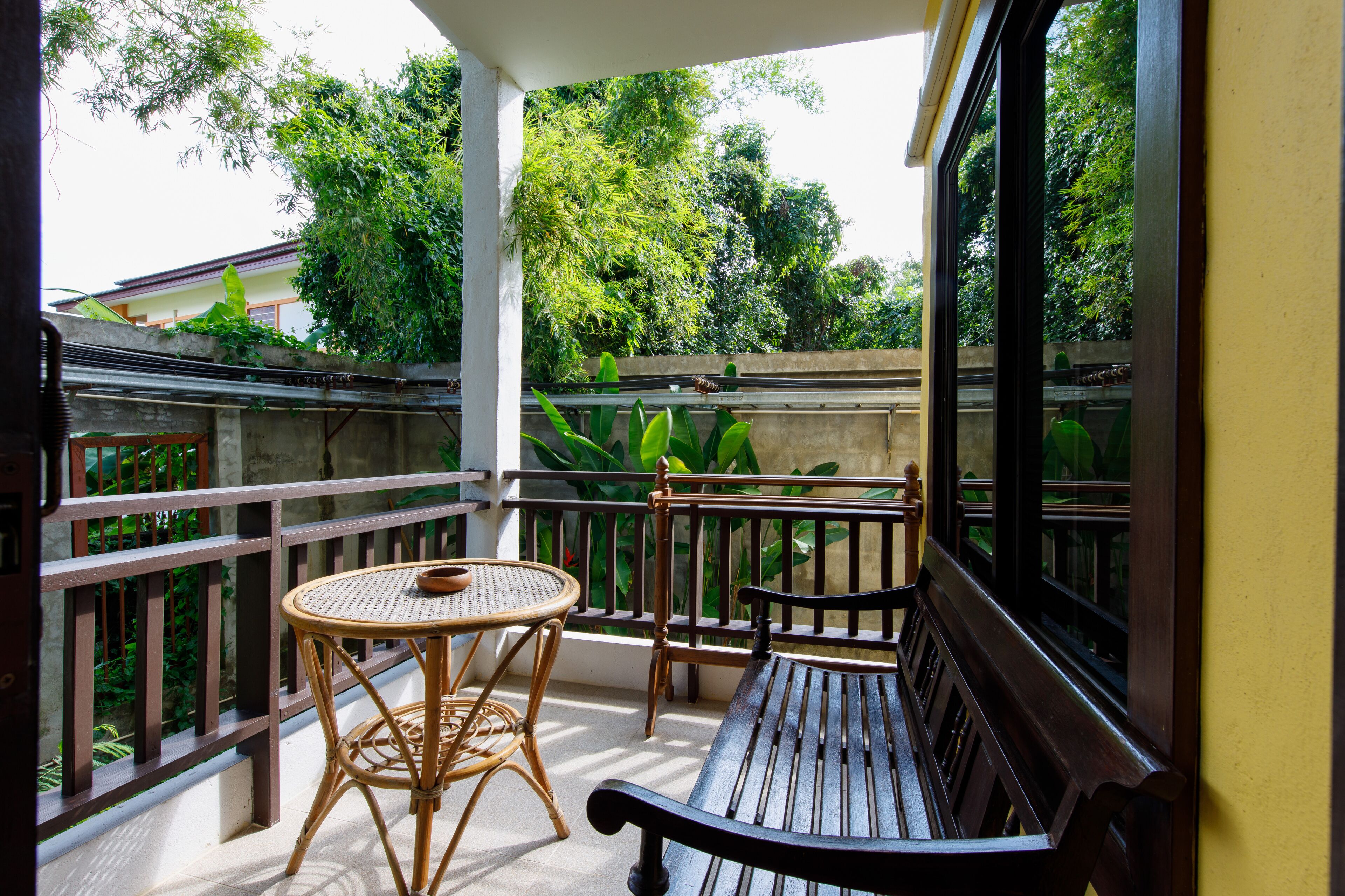 A balcony with a wooden bench and a wicker table, surrounded by greenery and a view of trees.