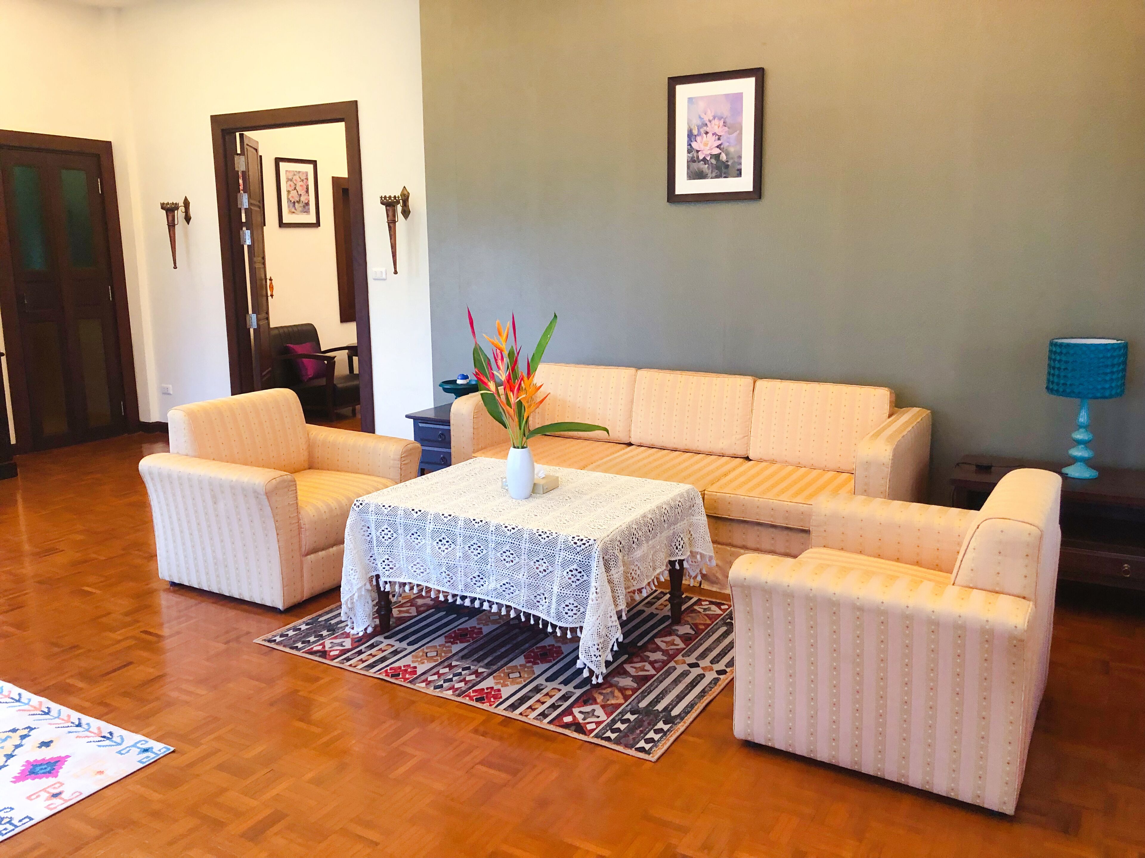 A living room with a beige sofa, two armchairs, a coffee table with a lace tablecloth, a vase with flowers, and a patterned rug.