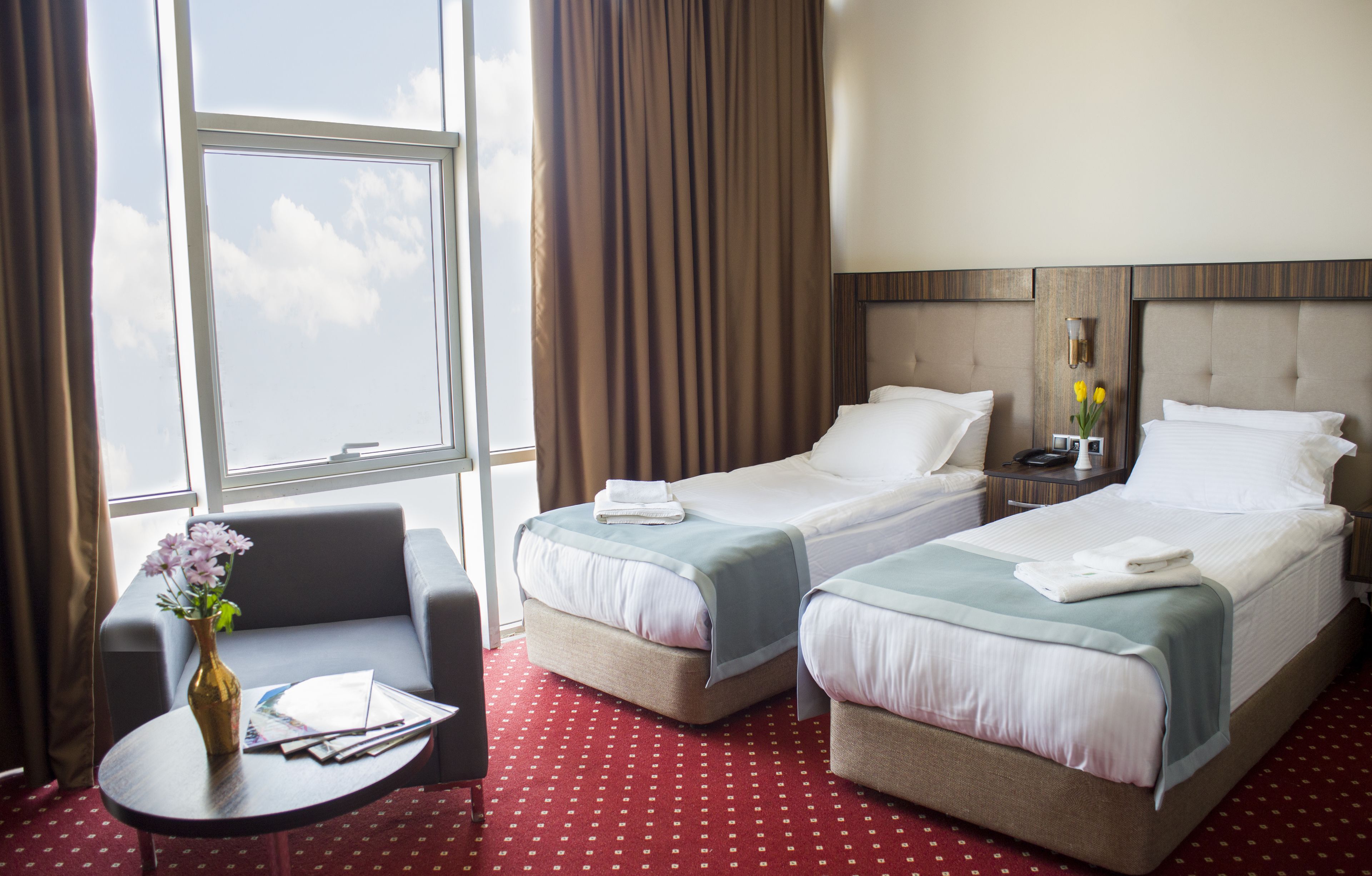 Habitación de hotel con dos camas, una mesita con un florero y vistas al cielo a través de amplios ventanales.