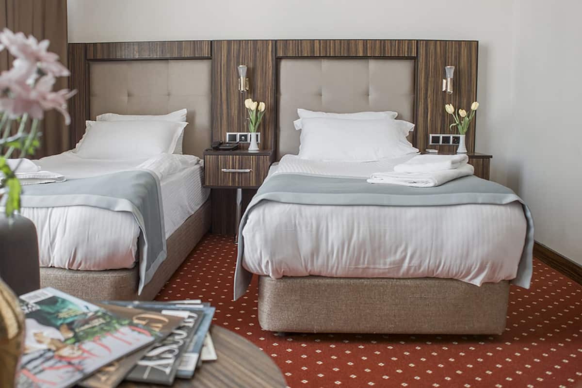 A hotel room with two beds, a small table with magazines, a vase with flowers, and a red patterned carpet.