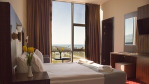 A hotel room with a large bed, a desk with a vase of flowers, a TV, and a view of the cityscape through large windows. - Legend Business Hotel Batumi (Batumi)