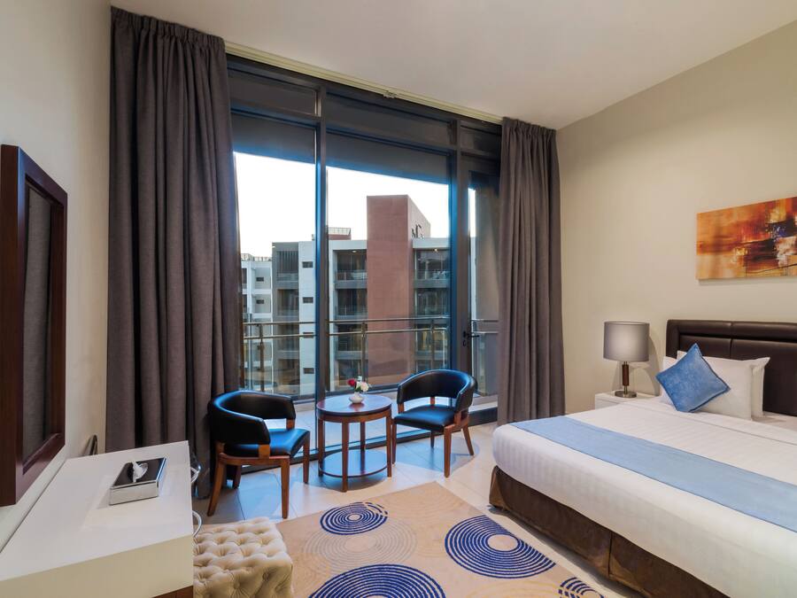 A hotel room with a large bed, a desk, two chairs, a small table, a lamp, and a painting on the wall.