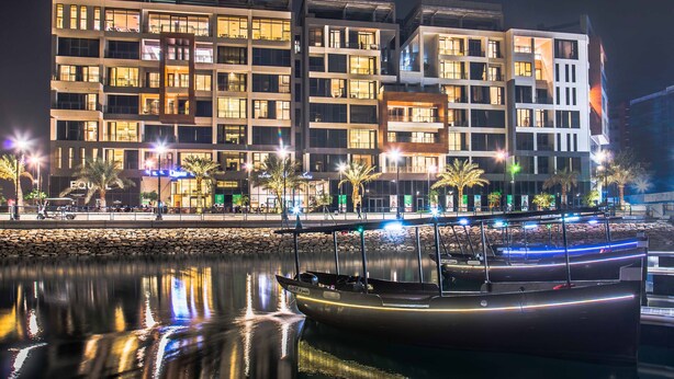 Un bâtiment moderne à plusieurs étages, doté de balcons, un port de plaisance avec des bateaux et des palmiers le long du littoral.