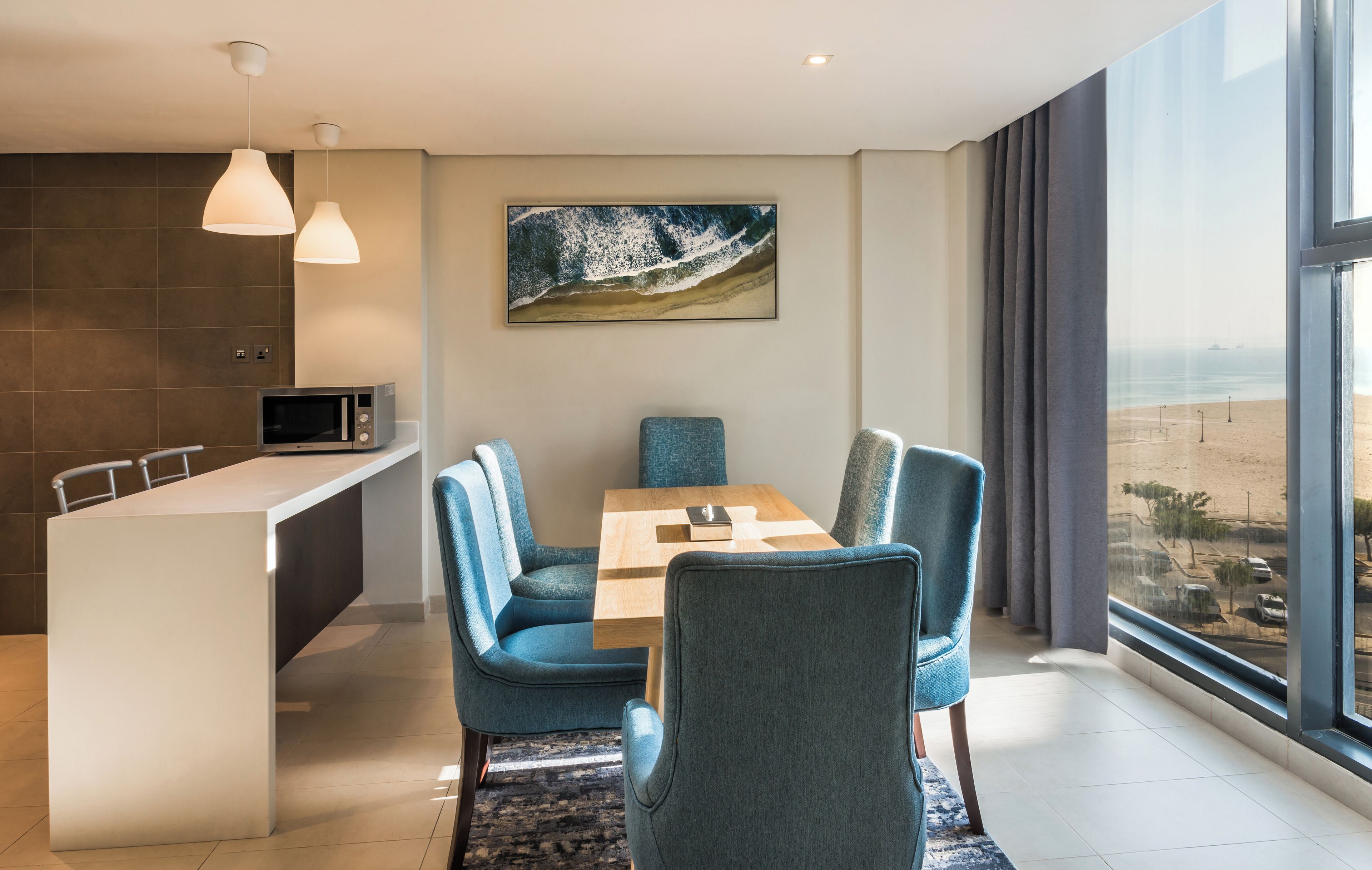 A modern hotel room with a dining area, a large window with a view of the beach, and a painting on the wall.