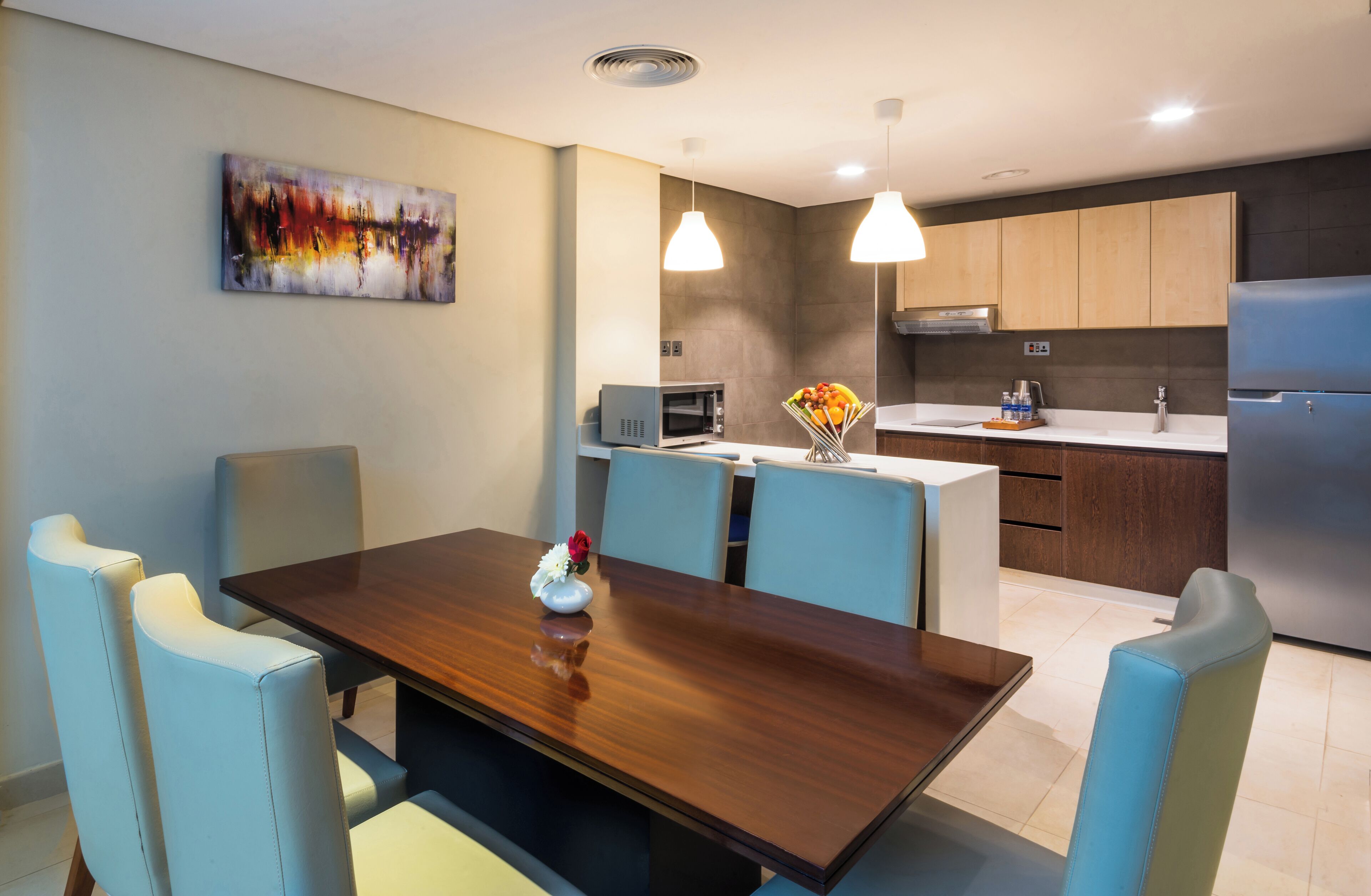 A modern kitchen with a central island, pendant lighting, and a dining area with a wooden table and cushioned chairs.
