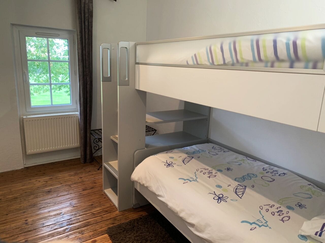 Una cama litera con colchón blanco y cabecero colorido. Se ve una ventana con vistas al exterior, con vegetación.