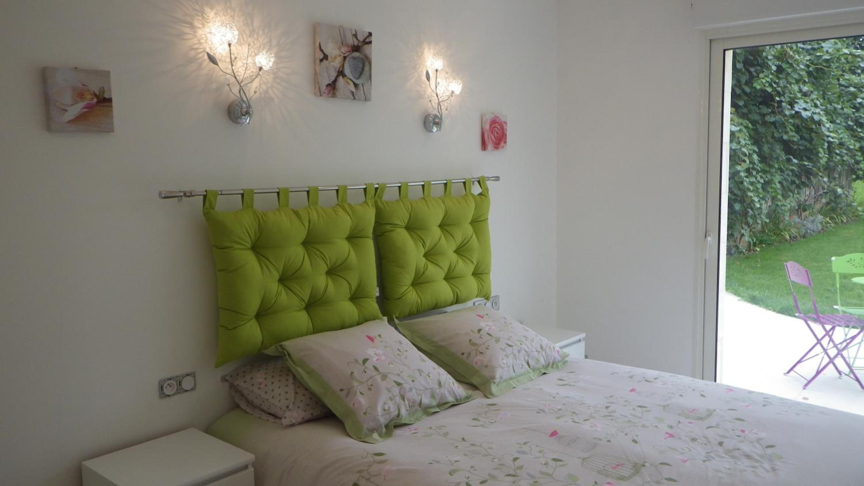 Un dormitorio con un cabecero verde con capitoné, ropa de cama floral y dos lámparas fijadas a la pared.
