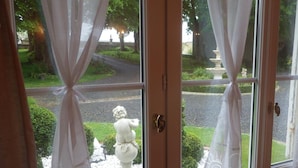 A view through a window with white curtains, a statue, and a garden with a fountain. - La Part des Anges (Rubercy)