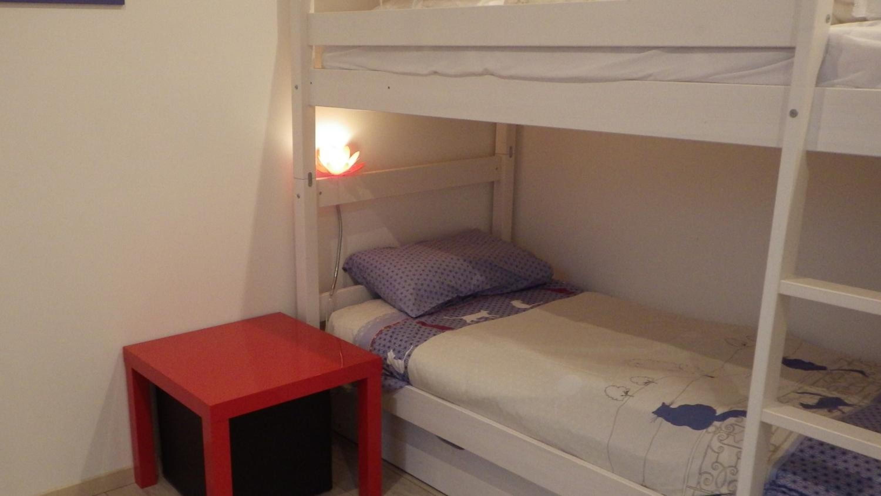 A bunk bed with a red table beside it, a single bed with a patterned blanket, and a wall-mounted shelf.