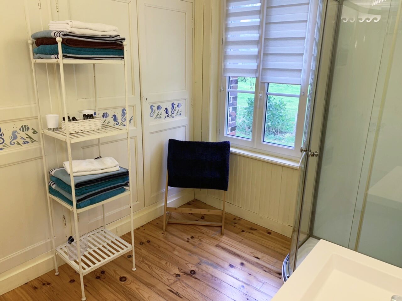 Une salle de bain avec un plancher en bois, un porte-serviettes blanc sur lequel sont pliées des serviettes, une chaise noire et un espace douche.