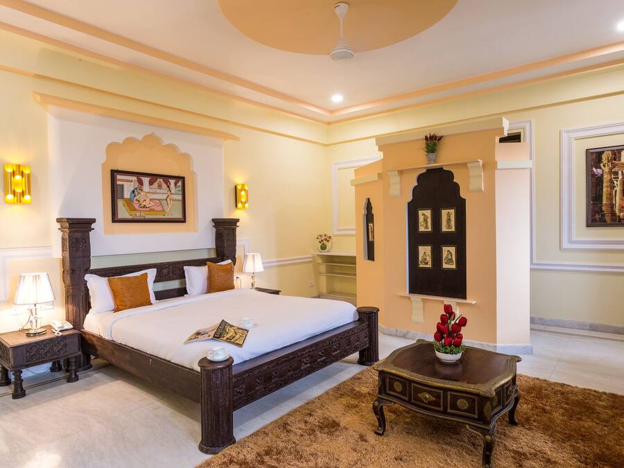 A well-lit bedroom with a large bed, bedside tables, a nightstand, a decorative mirror, and a small table with a vase of flowers.