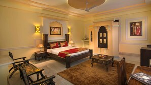 A bedroom with a bed, bedside tables, a chair, a small table, and a ceiling fan. - Syna Heritage Hotel (Rajnagar)