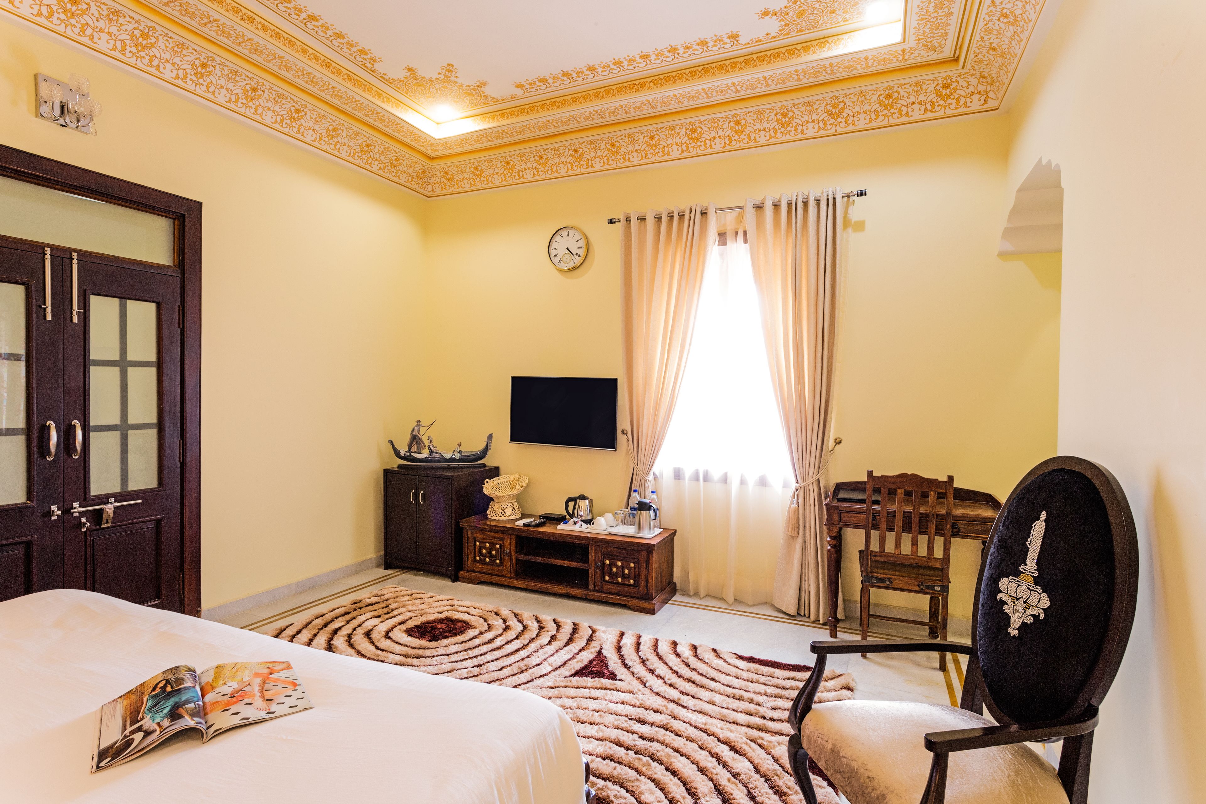 Une chambre d’hôtel comprenant un lit, une télévision, une chaise en bois, un bureau avec une lampe et un plafond décoratif.
