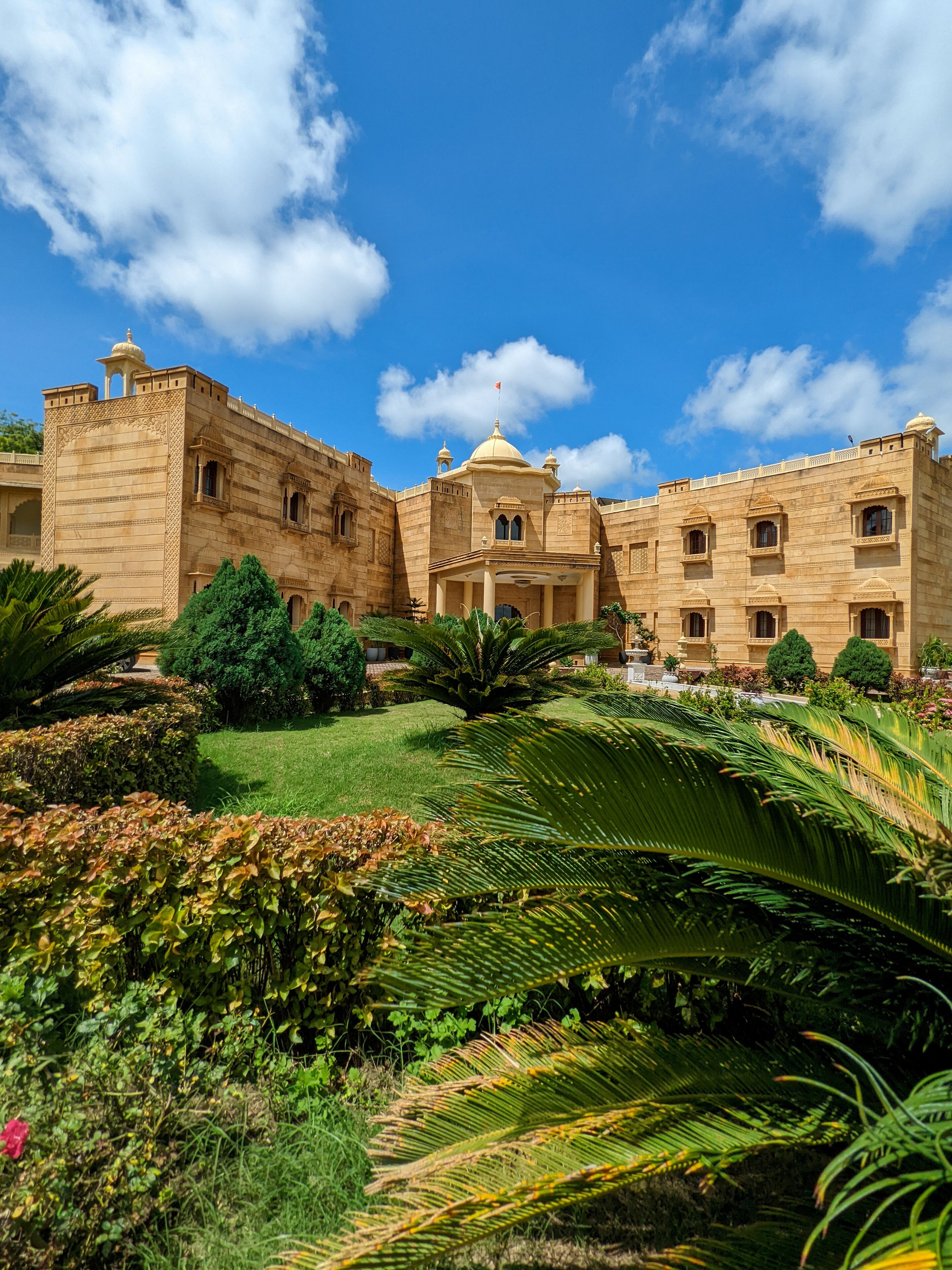 A large, historic building with intricate architecture, surrounded by lush gardens and a clear blue sky.
