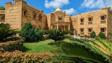 A large, historic building with intricate architecture, surrounded by lush gardens and a clear blue sky.