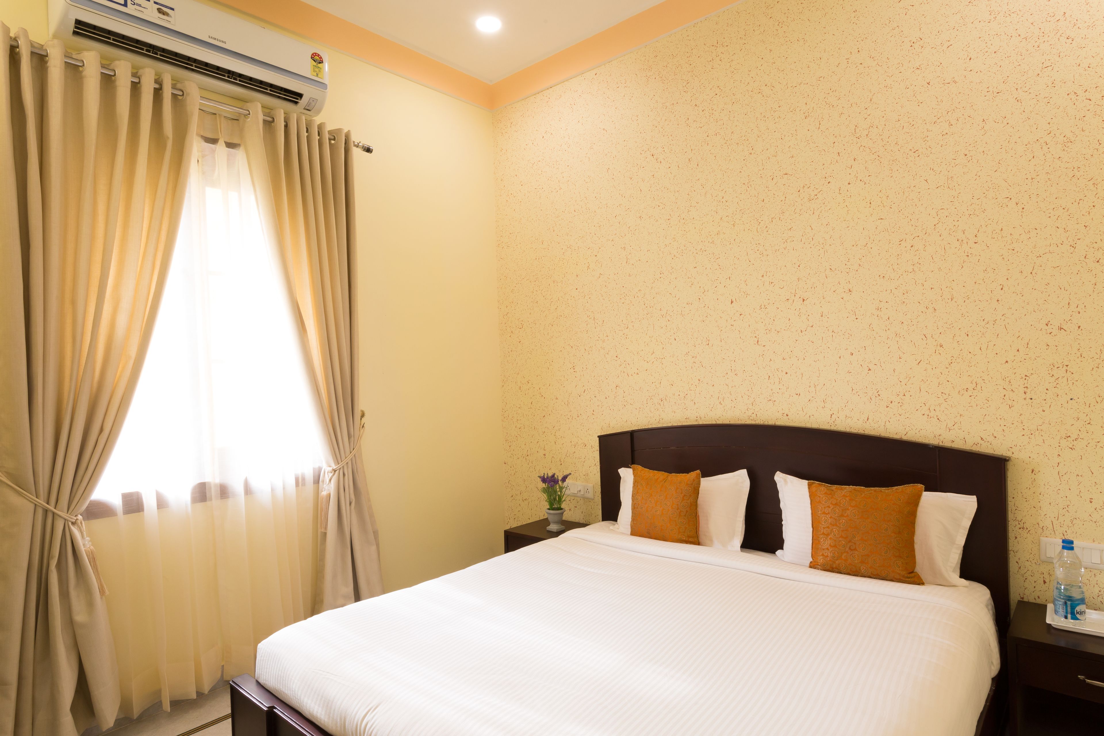 Una camera d'albergo con un letto grande, tende color beige, un condizionatore d'aria, un comodino con un vaso di fiori e una parete con un motivo screziato.