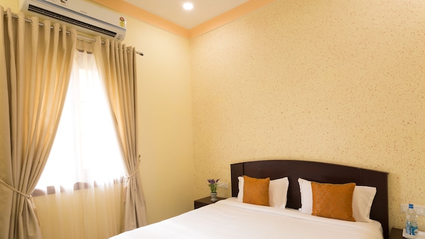 A hotel room with a large bed, beige curtains, an air conditioning unit, a nightstand with a vase of flowers, and a wall with a speckled pattern. - Syna Heritage Hotel (Rajnagar)