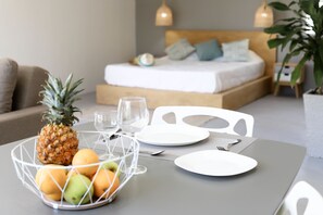 Una mesa de desayuno ordenada con frutero, platos y cubiertos. Se ve una cama con cabecera de madera y almohadas al fondo.