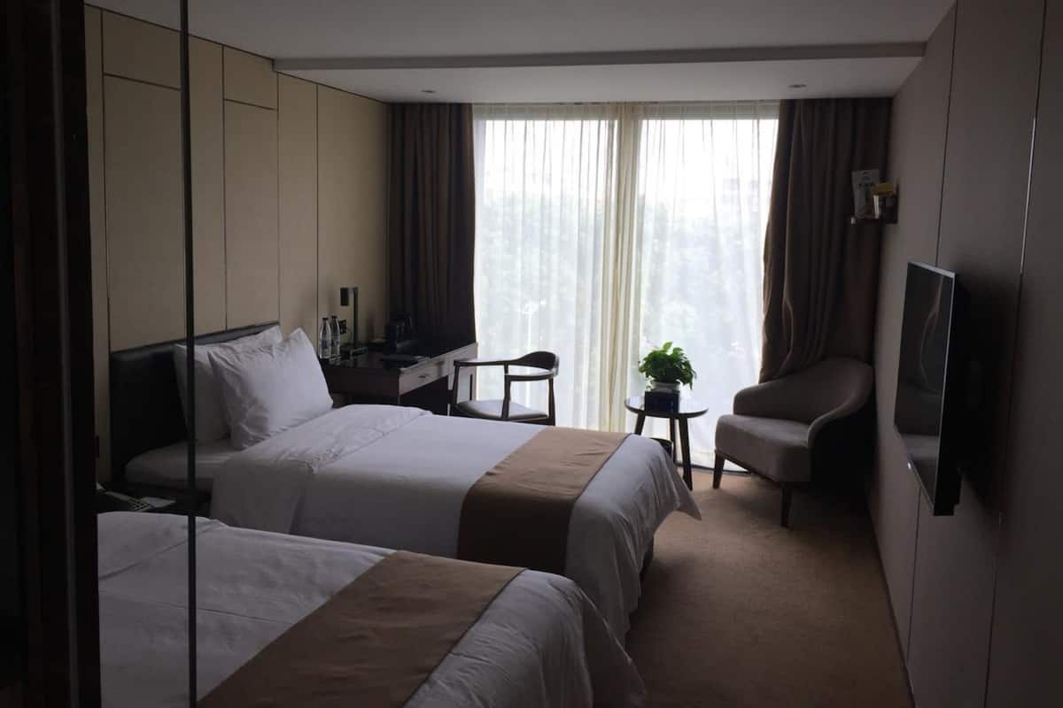 Quarto de hotel com duas camas, uma escrivaninha com cadeira, televisão e janela com cortinas.