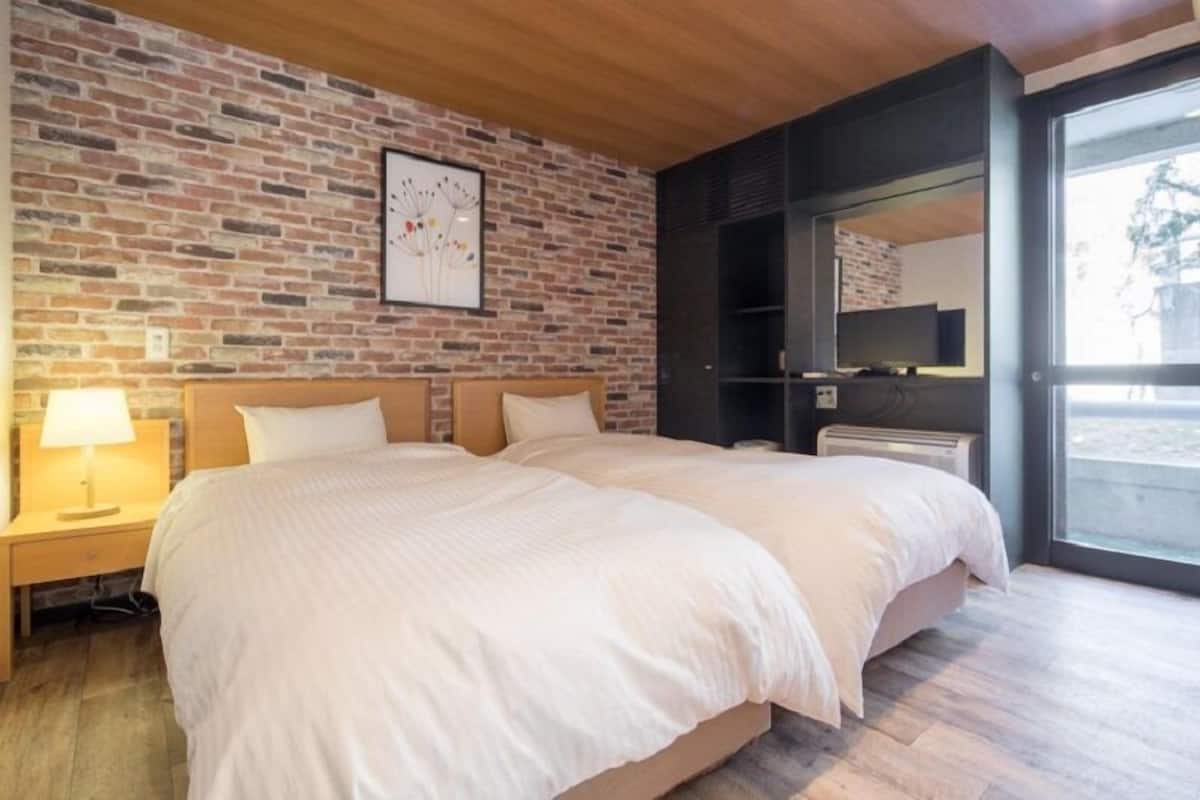 Una cama doble con cabecera de madera, una mesita de noche con lámpara, pared con diseño de ladrillo, un espejo grande y vistas al exterior a través de una puerta de cristal.