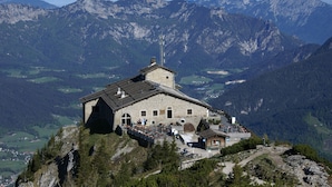 A mountain lodge with a stone facade, a sloped roof, and a small annex. It is situated on a rocky outcrop with a view of the surrounding mountains.