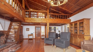 A spacious loft-style room with a high ceiling, wooden beams, a staircase, and a view of a forest path through a large window.