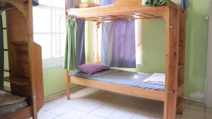 A bunk bed with a wooden frame, a mattress, and a pillow. There is a window with curtains, and a wall with a light switch. - Lotus Garden Hostel (Izmir)