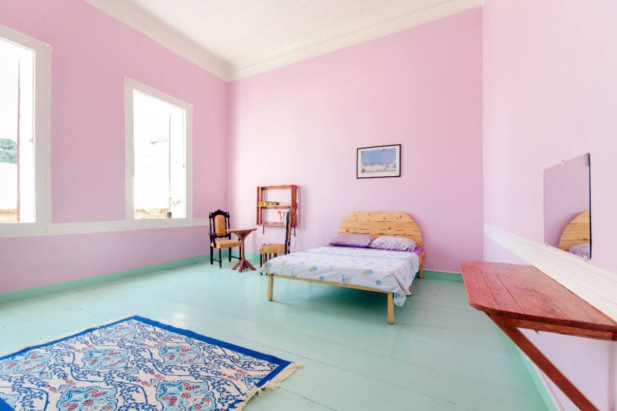 A pink-walled bedroom with a bed, a wooden nightstand, a chair, a small table, and a large window.