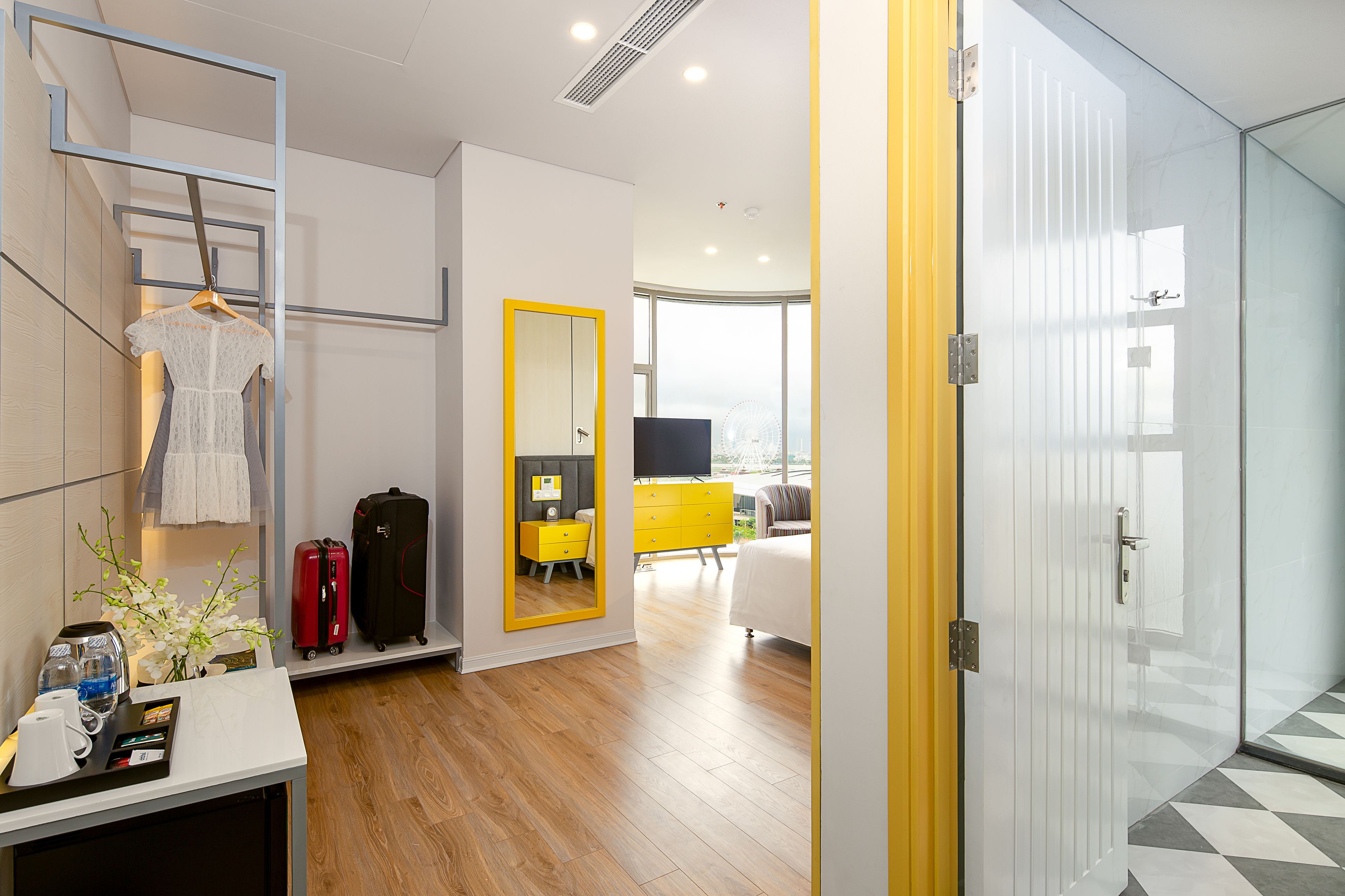 A hotel room with a wooden floor, a mirror with a yellow frame, a bed, a yellow dresser, and a white wall with a window.