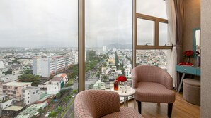 Ein Hotelzimmer in einem Hochhaus mit Blick auf die Stadt, zwei karierte Sessel, ein kleiner Tisch mit einer Vase roter Blumen und ein blauer Schreibtisch mit Spiegel.