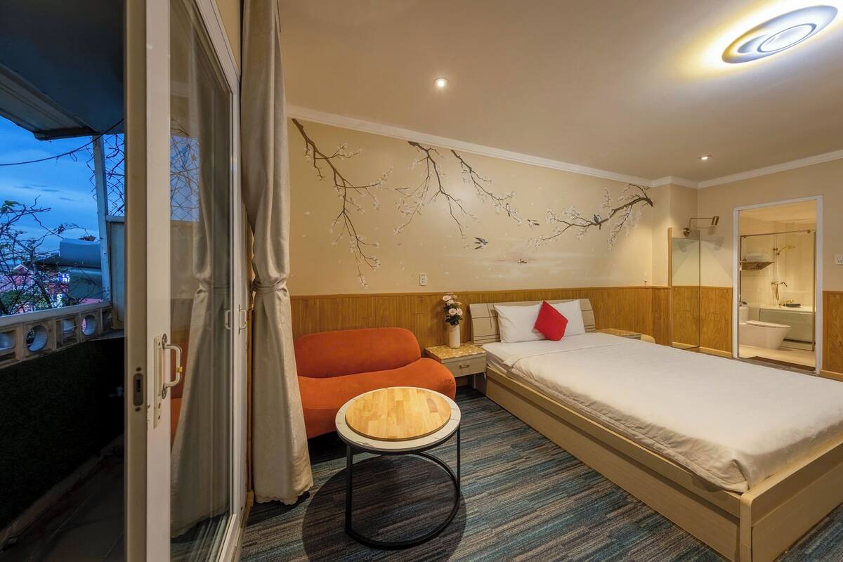 Habitación de hotel con cama, una mesita, una mesita de noche, una ventana con cortinas y una obra de arte colgada en la pared.