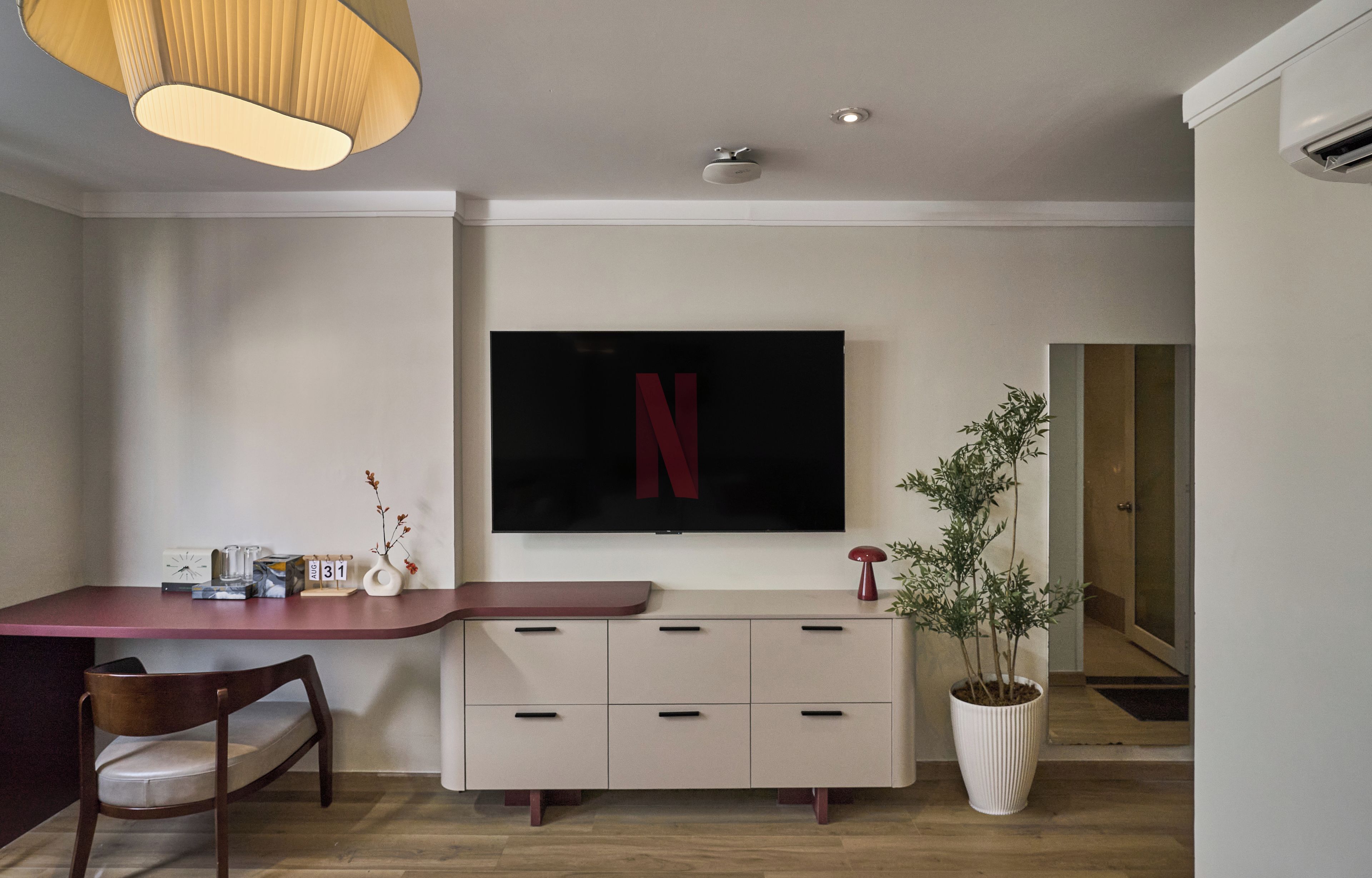 Ein modernes Wohnzimmer mit einem wandmontierten Flachbildfernseher, einem Holzessttisch mit Stühlen, einer Topfpflanze und einer eingebauten Kommode mit Schubladen.