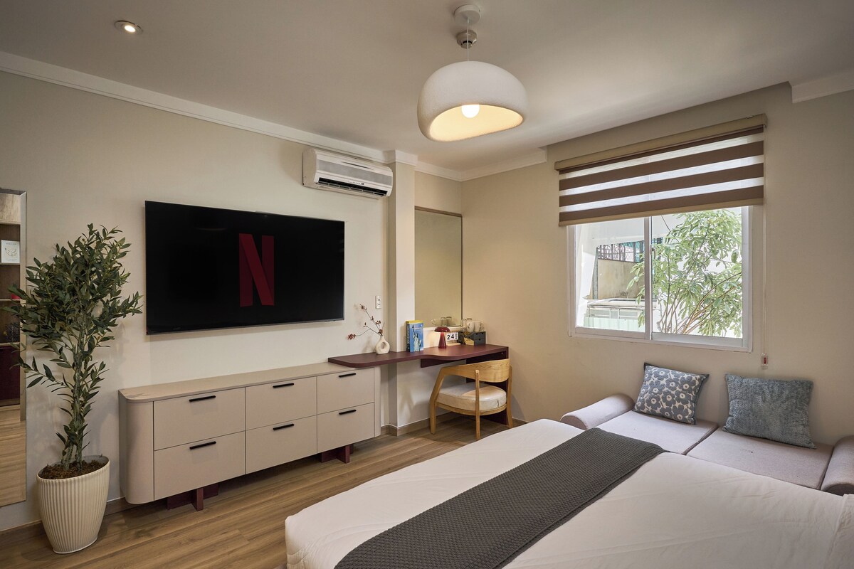 A modern bedroom with a flat-screen TV, a desk, a chair, a bed, and a large window.