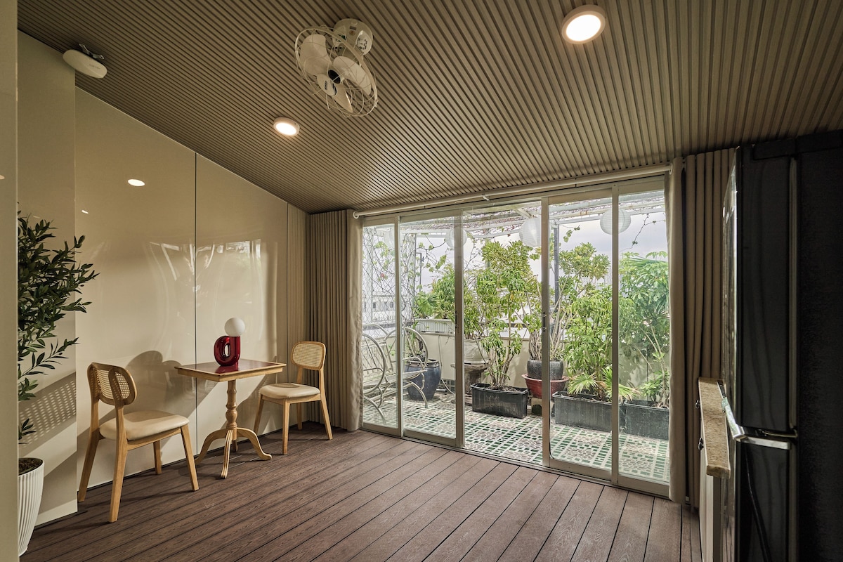 A room with a wooden floor, a table, two chairs, a potted plant, and a sliding glass door leading to an outdoor area with plants and a view of the sky.