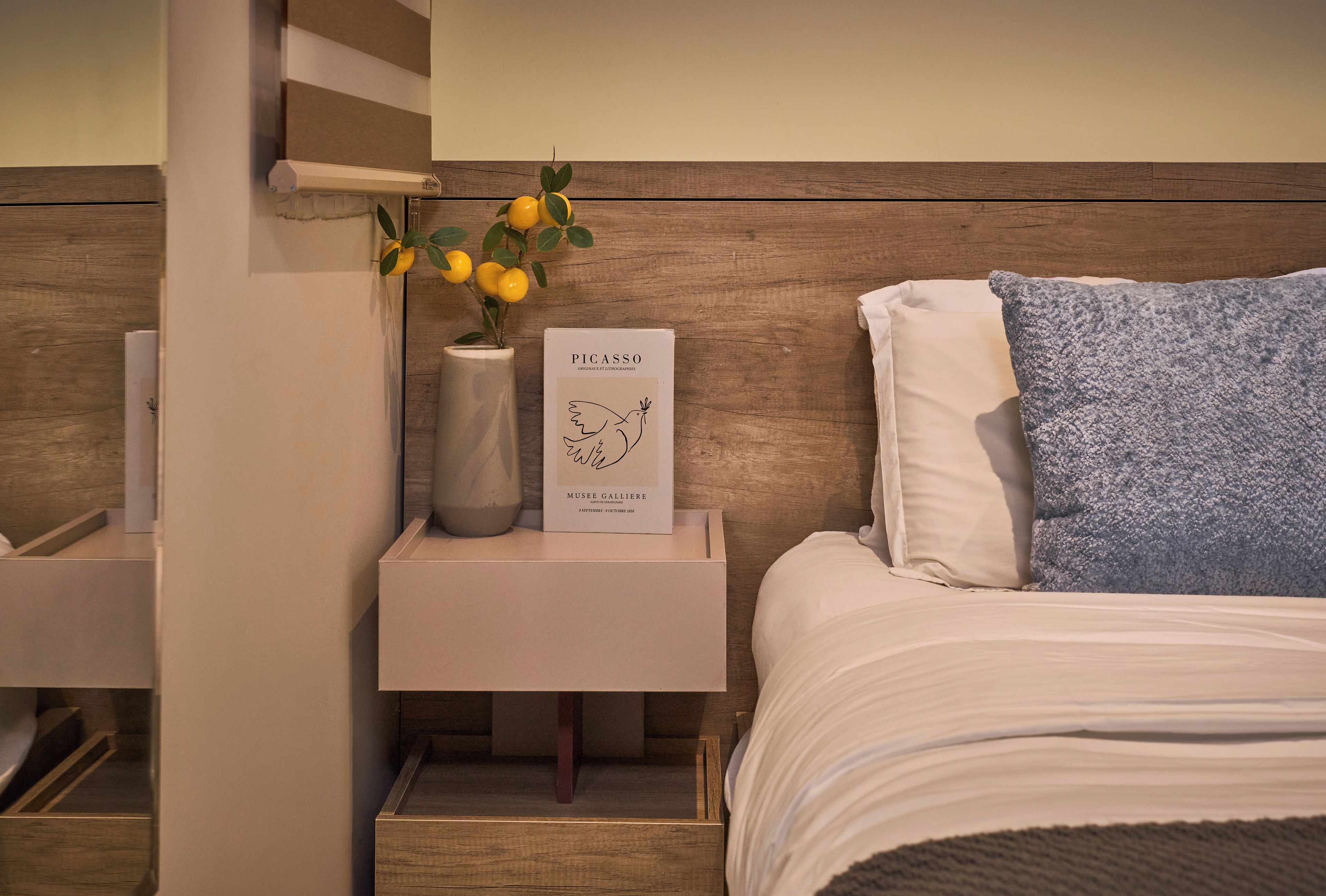 Un dormitorio ordenado con una cama, una mesita de noche con un jarrón de flores y un libro titulado 'PICASSO'.