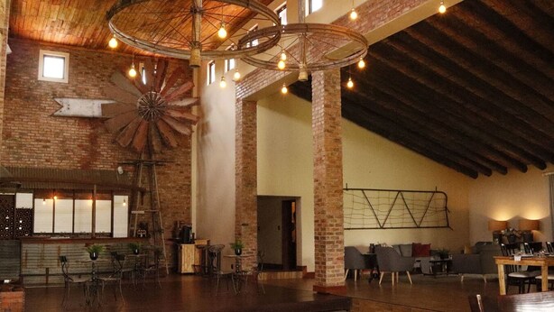 A spacious interior with exposed brick walls, a large wooden ceiling, and a central windmill-like structure. There are multiple seating areas with tables and chairs, and a bar area with a rustic design. - Water on Waldrift (Vereeniging)