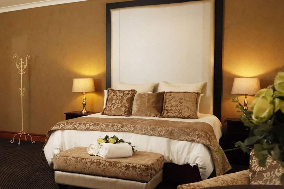 Hotelkamer met een groot bed, twee bedlampen, een decoratieve spiegel en een klein tafeltje met een handdoek en bloemen.