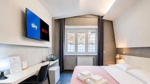 A hotel room with a bed, a desk, a chair, a TV displaying Sky and Netflix logos, and a window with curtains. - Le Vie del Vaticano (Rome)