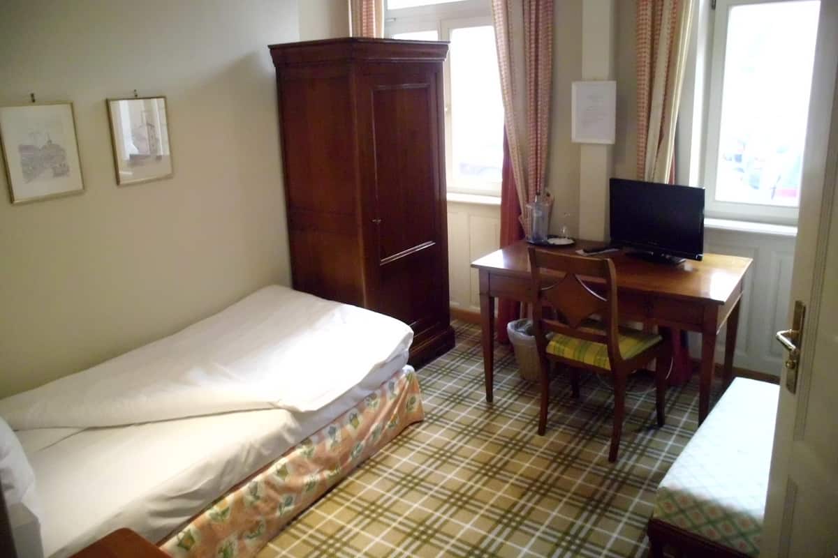 A small, simple room with a bed, a desk with a chair, a wardrobe, and a television.