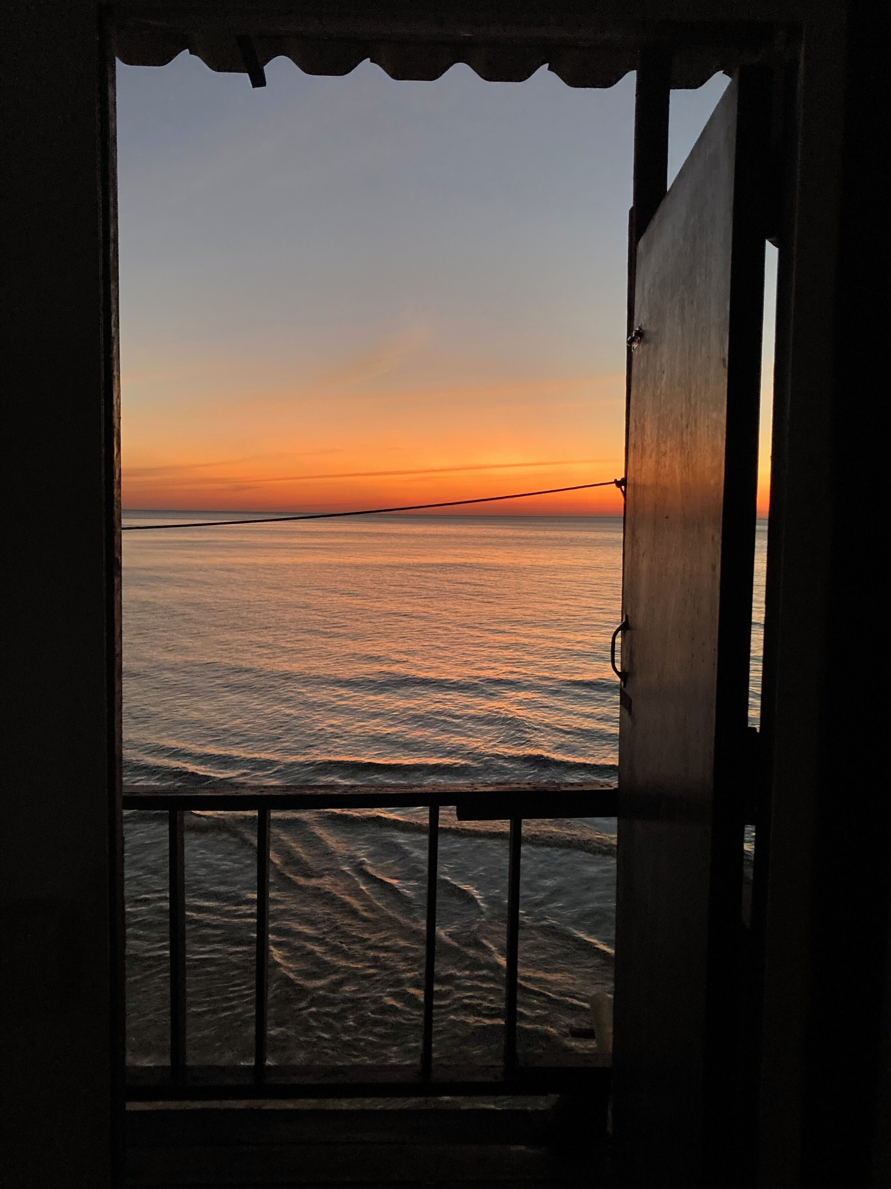 A view through an open door with a railing, overlooking a calm sea at sunset.