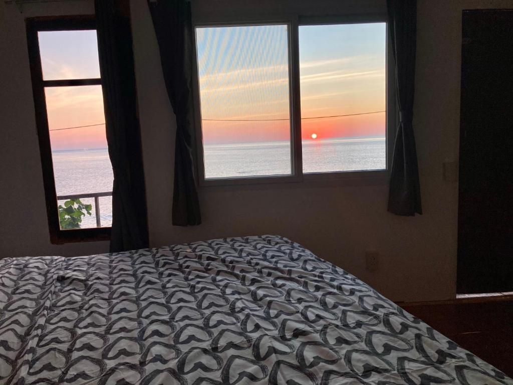 Una habitación con cama, dos ventanas y vistas a la puesta de sol sobre el mar.